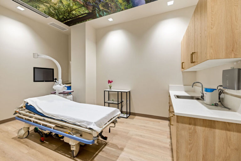 A modern, clean medical examination room with a hospital bed, medical equipment including a monitor and overhead lamp, wood cabinetry, a sink, and a small table with a vase of flowers; the ceiling features an image of green foliage.