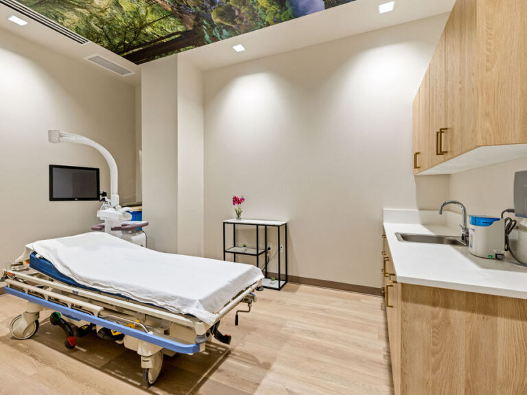 A modern, clean medical examination room with a hospital bed, medical equipment including a monitor and overhead lamp, wood cabinetry, a sink, and a small table with a vase of flowers; the ceiling features an image of green foliage.