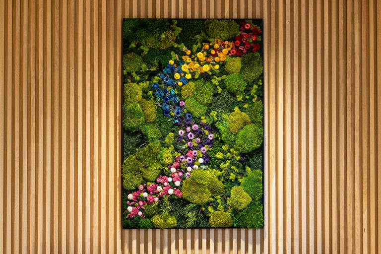 A vertical rectangular frame filled with green moss, various plants, and clusters of colorful flowers arranged in a diagonal, wavy pattern, mounted on a wall with vertical wooden slats.