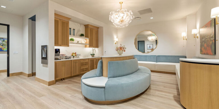 Modern waiting room with light wood flooring, soft blue and white seating, built-in cabinetry, a decorative chandelier, a round mirror, and artwork on the walls.
