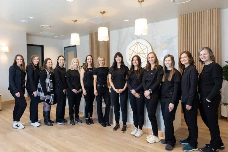 Group of thirteen Lyra team members standing in a modern, well-lit office space with wood floors and pendant lights, most wearing coordinated black outfits, smiling and posing for a group photo in front of a reception desk with a geometric gold logo on the wall.