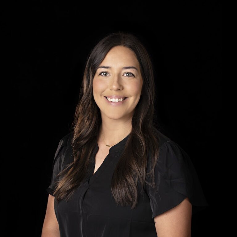Lyra team member Ashley Knollin smiling, wearing a black blouse, and posed against a black background.