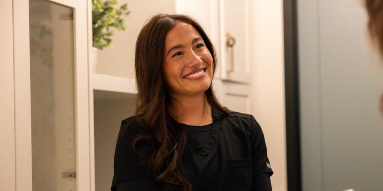 Smiling woman in black scrubs interacting warmly with another person, possibly a patient, in an indoor professional or medical setting.