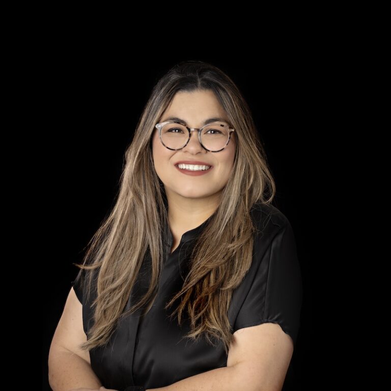 Lyra team member Monique Manansala with long, light brown hair wearing glasses and a black shirt, smiling against a plain black background.