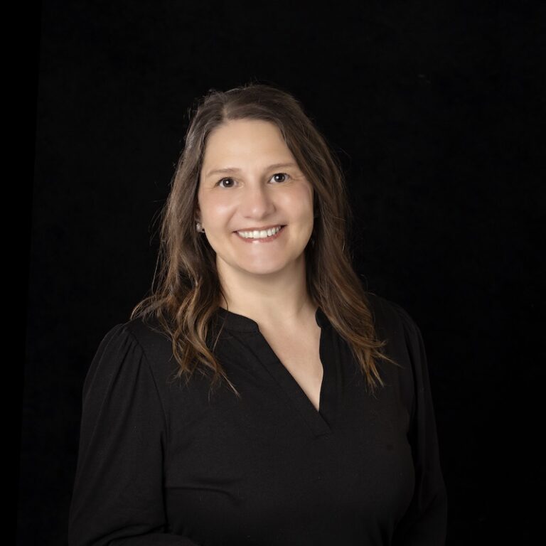 Lyra team member Jessica Strong smiling with long brown hair wearing a black blouse, posed against a plain black background.