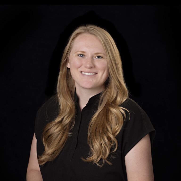 Lyra team member Myesha, smiling and wearing a black shirt, with long wavy blonde hair, photographed against a black background.