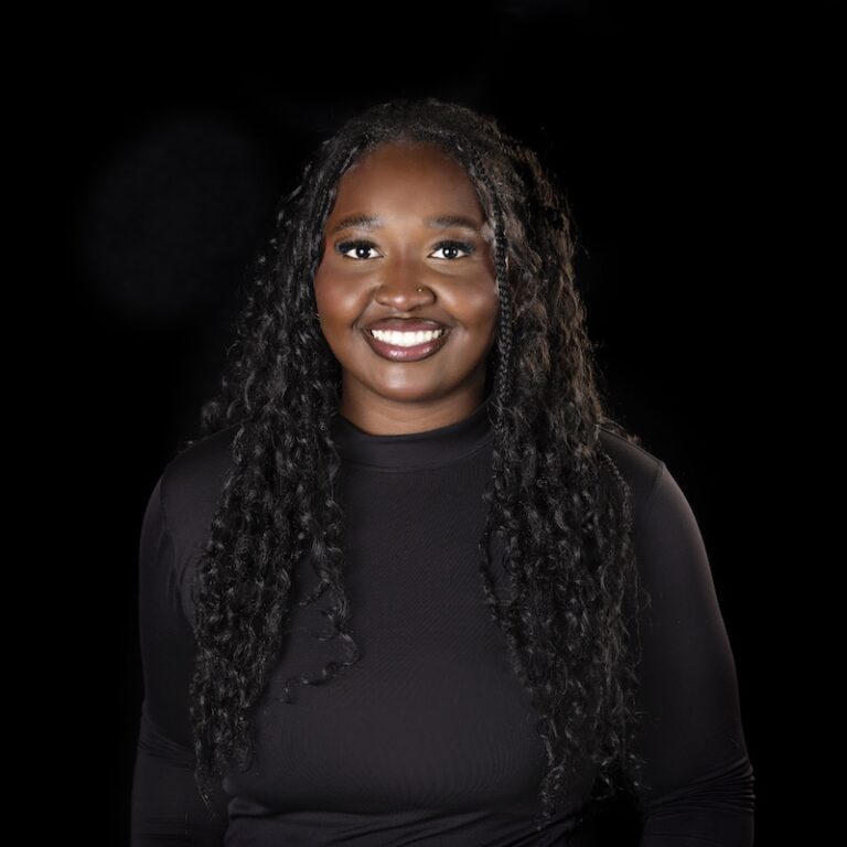 Smiling woman with long curly hair, wearing a black long-sleeve top, posed against a black background.