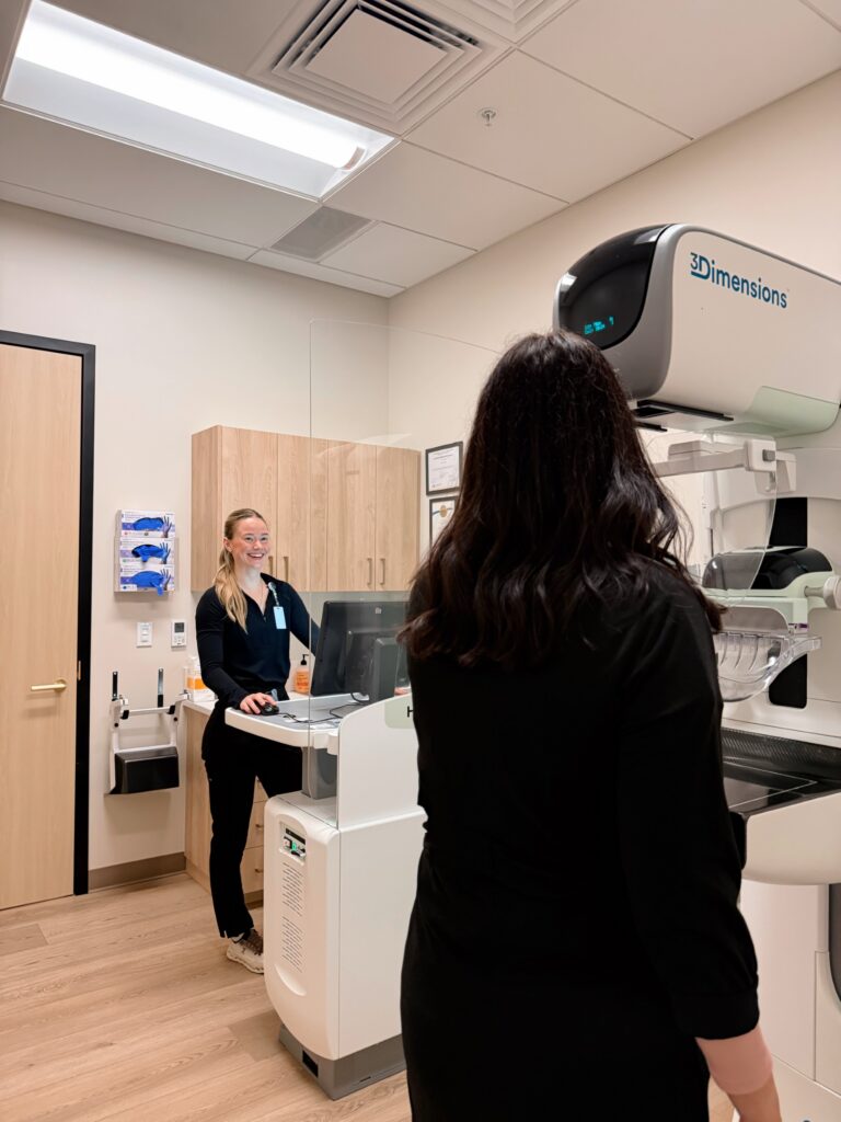 A healthcare professional prepares a mammogram machine while a woman stands in position for breast imaging in a modern medical exam room.