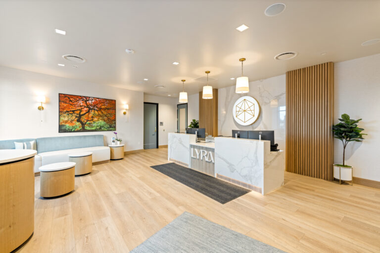 Modern office reception area with marble desk, light wood flooring, decorative lighting, seating, a large wall art, and a potted plant.