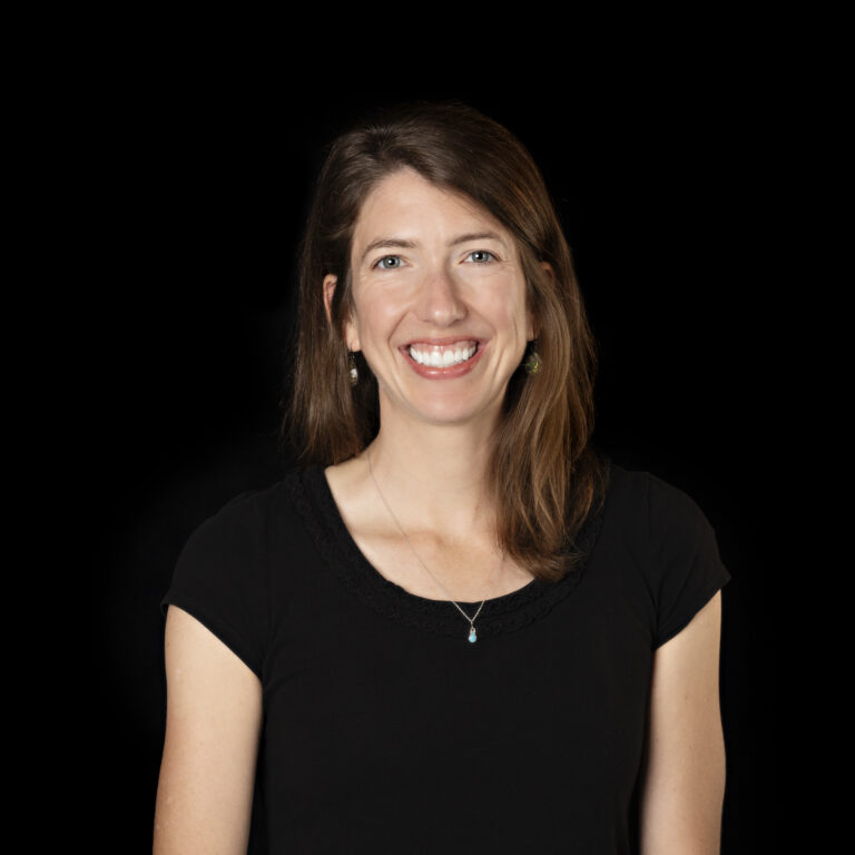 Woman with long brown hair wearing a black top, smiling against a plain black background.