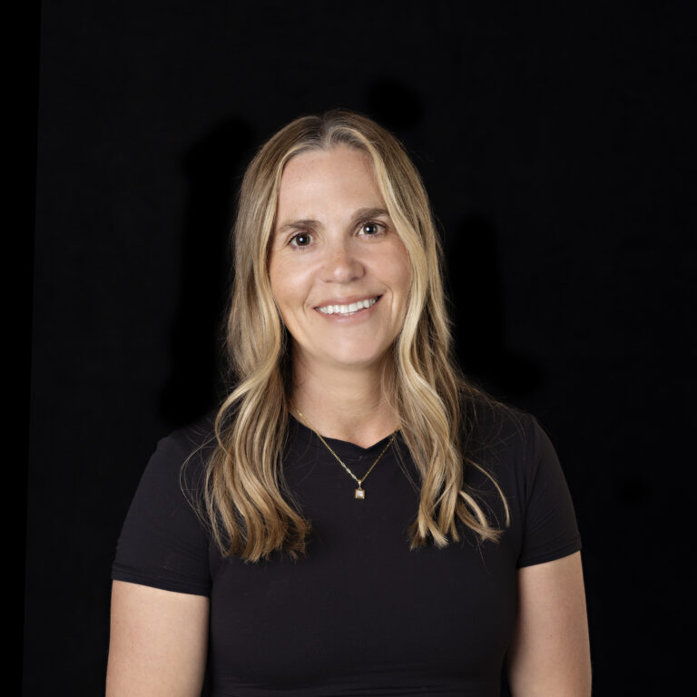 Woman with long blonde hair wearing a black short-sleeve shirt and a gold necklace, smiling against a plain black background.