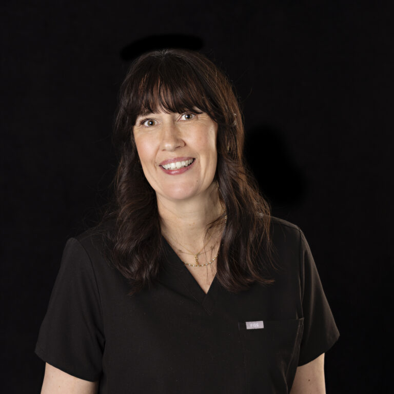 Woman with wavy dark hair and bangs, wearing a black top and gold necklaces, smiling against a black background.