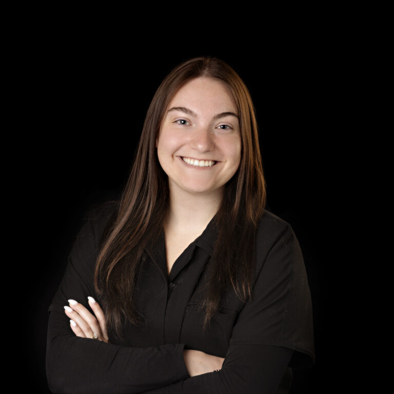 A young woman with long straight brown hair, wearing a black shirt, smiling with her arms crossed against a black background.
