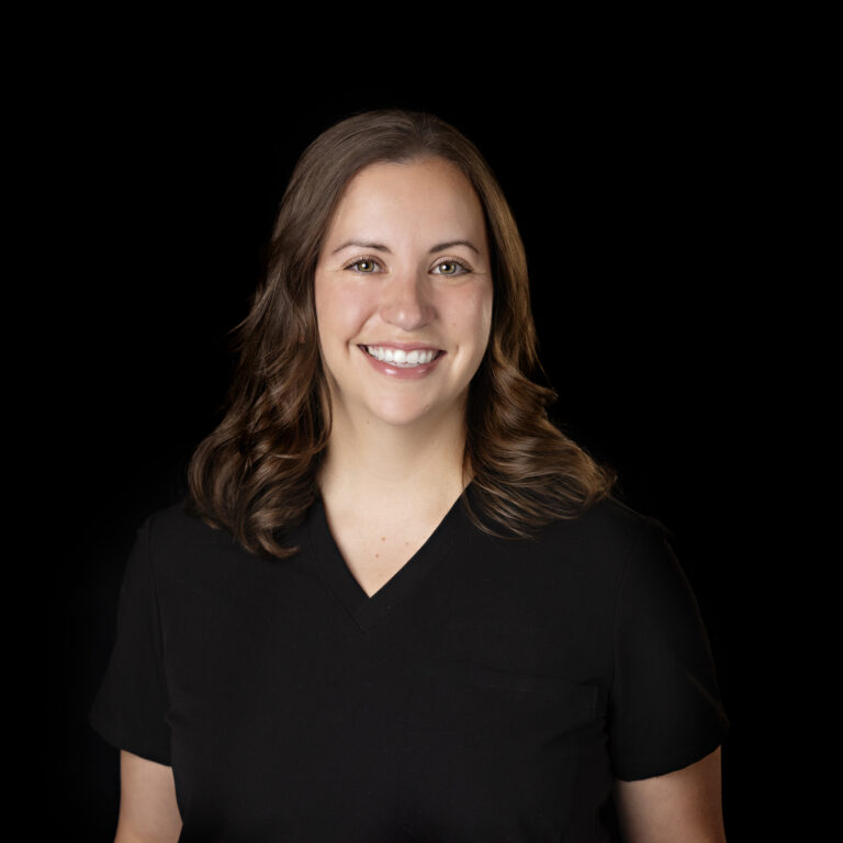 Smiling woman with medium-length brown hair, wearing a black V-neck top, posed against a plain black background.