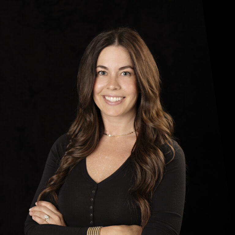 Woman with long wavy brown hair wearing a black top, smiling with arms crossed against a black background.